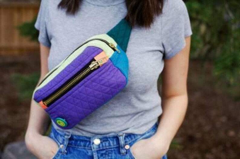 K&ouml;nnte beinhalten: Eine mehrfarbige gesteppte Bauchtasche mit Rei&szlig;verschluss und einem kleinen runden Knopf. Die Bauchtasche besteht aus verschiedenen Stoffen, darunter Lila, Blau, Gr&uuml;n und Gelb.