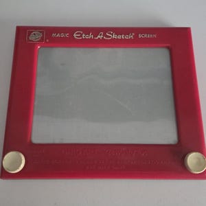 May include: A red plastic drawing toy with a rectangular frame and a gray screen. The top of the frame features the text MAGIC ETCH A SKETCH SCREEN. Two white circular knobs are positioned at the bottom left and bottom right corners for control.