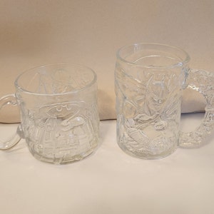 May include: Two clear glass mugs with a Batman design. The mugs have a handle and are embossed with a Batman character.