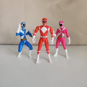 May include: Three Power Ranger action figures in a row. The figures are in blue, red, and pink, each with a white diamond-shaped design on the chest. They have white boots and gloves, and helmets with visors.