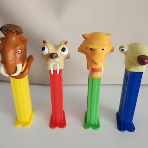 May include: Four PEZ dispensers featuring characters from the animated film "Ice Age." The dispensers have colorful plastic bases and character heads, including a mammoth, a saber-toothed tiger, and a sloth. The dispensers are yellow, red, green, and blue.