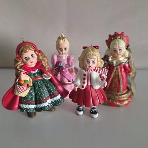 May include: Four decorative figurines featuring dolls in distinct outfits: one in a green dress with a red cape and basket, one in a pink gown, one in a red skirt with a white cardigan holding a treat, and one in a red and gold ornate dress.