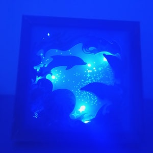 Op de afbeelding: Een vierkante, verlichte schaduwdoos met een dolfijnontwerp. Het kunstwerk is verlicht met blauwe lichten, waardoor een gloeiend effect ontstaat. Het ontwerp bevat dolfijn silhouetten en sterachtige accenten.