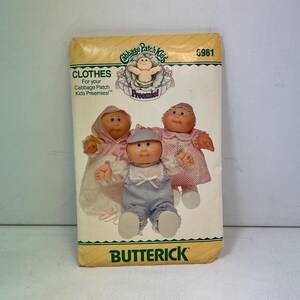 May include: A vintage Cabbage Patch Kids clothing pattern packet. The packet features illustrations of three Cabbage Patch Kids dolls wearing various outfits. The packet has the words "CLOTHES" and "BUTTERICK" printed on it.