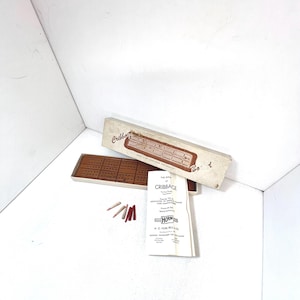 May include: A wooden cribbage board game with a brown wooden board and white pegs. The game is in its original box with the rules of cribbage printed on a white paper. The box is labeled "The Game of Cribbage" and has a red and white logo for Horn.
