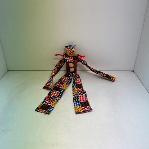 May include: A handmade wooden doll with a painted face, wearing a patchwork fabric outfit. The doll has a rectangular body, two white button accents, and arms and legs made of the same colorful fabric. A small hat and red yarn accents complete the design.