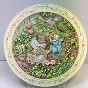 May include: A decorative ceramic plate featuring a relief scene of cats and a mouse in a garden setting. The plate has a cream-colored border with pink roses and green vines. The scene includes pink roses, blue flowers, and lush green foliage.