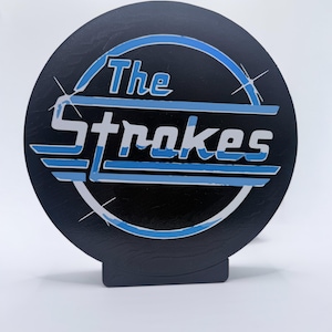May include: A black circular stand-up sign featuring the band name "The Strokes" in white and blue lettering. The design includes a blue arc and lines, with starburst accents. The sign is on a white background.