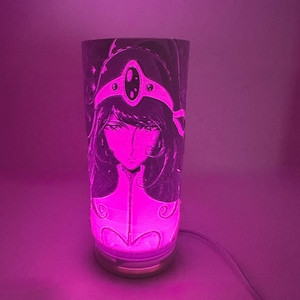 May include: A cylindrical lamp with a detailed illustration of a woman's face and a decorative headpiece. The lamp emits a bright pink glow, and the base has a gold finish. A power cord is attached.