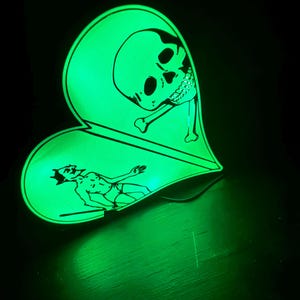 May include: A glowing green heart-shaped decorative item with a black outline. The design features a skull and crossbones on one side and a reclining figure on the other. The item is illuminated against a dark background.