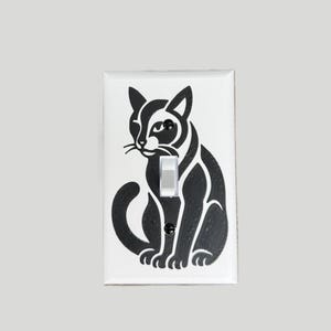 May include: A white light switch cover featuring a black silhouette of a cat in a seated position. The cat has pointed ears, a curved tail, and a detailed facial expression. The switch is a standard toggle style.