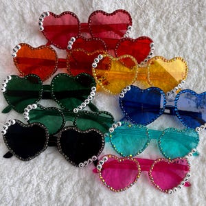 Puede incluir: Un conjunto de diez gafas de sol en forma de corazón con pedrería y monturas de diferentes colores. Cada par tiene un nombre diferente escrito en cuentas blancas en la montura. Los nombres son: Bang Chan, Lee Know, Changbin, Hyunjin, Han, Felix, Seungmin, I.N, Jeongin y Seungmin.