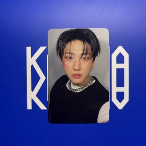 May include: A portrait card featuring a person with dark hair, wearing a black vest and a silver chain necklace. The person has makeup with star accents on their face. The background is a solid blue with white geometric shapes.