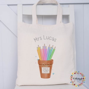 May include: Cream-colored tote bag with the name "Mrs Lucas" printed in gray. The bag features a watercolor illustration of colored pencils in a terracotta pot with a tag that reads "Thank you for helping me grow."