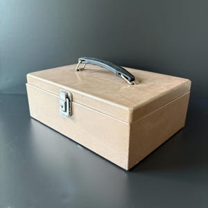 May include: A tan metal toolbox with a black handle and a silver latch. The rectangular box is closed and sits on a dark surface. The handle is centered on the top of the box.