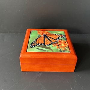 May include: A small wooden box with a hinged lid. The lid features a colorful image of a monarch butterfly on a green background with orange flowers.