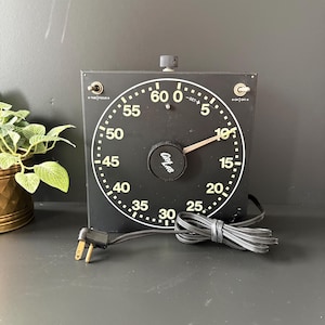May include: A black square GraLab darkroom timer with a large circular dial featuring white numbers from 0 to 60. The device includes a central black knob, two metal toggle switches at the top corners, and a black electrical power cord.