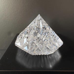 May include: A clear, faceted crystal pyramid with a pointed top. The pyramid is made of clear glass and has a geometric design. The pyramid is sitting on a black surface.