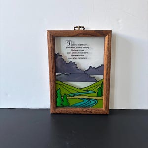 May include: A framed artwork with a landscape scene. The artwork features a wooden frame, a glass panel, and a painted landscape with green hills, a blue river, and dark gray clouds. The text reads: "I believe in the sun..."