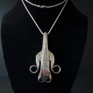 May include: A silver pendant necklace featuring a repurposed fork design. The fork's handle is adorned with floral patterns, and the tines are shaped into a unique, artistic form. The necklace hangs on a delicate silver chain, creating a vintage-inspired accessory.