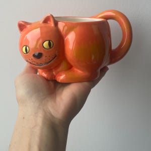 May include: Orange ceramic cat mug with a wide, toothy grin and yellow eyes. The mug has a handle shaped like a cat's tail.