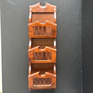 May include: A vintage wooden wall-mounted organizer with three tiered compartments. The organizer is made of dark brown wood and features a decorative top with a curved cutout. Each compartment has a rectangular opening with vertical slats, ideal for storing mail or documents.