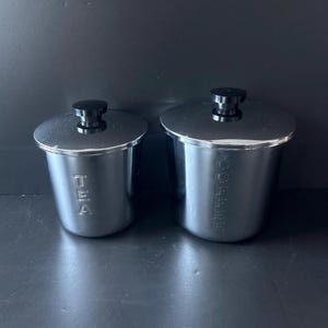 May include: Two stainless steel canisters with black lids. One canister is labeled "TEA" and the other is labeled "COFFEE". The canisters are cylindrical with a shiny, reflective surface. The lids have a small, black knob.