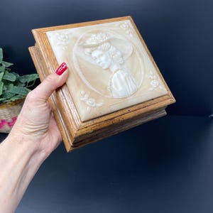 May include: A wooden box with a raised, white cameo of a woman wearing a hat. The cameo is set in a light brown background with floral accents.