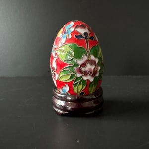 May include: A decorative, egg-shaped ornament with a red background, featuring a floral design in green, white, and purple. The egg sits on a dark brown wooden stand. The floral design is outlined in gold.