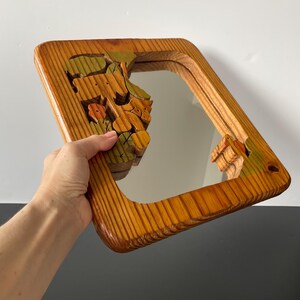 May include: A wooden mirror with a carved frame featuring a colorful floral design. The mirror is surrounded by a thick wooden frame with a natural wood finish.