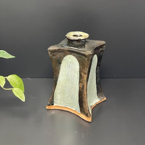 May include: A small, square, ceramic vase with a brown glaze and white accents. The vase has a slightly flared opening and a rounded base.