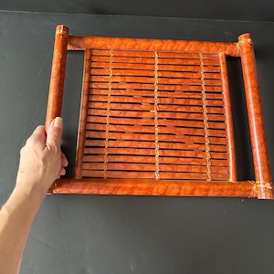 May include: A rectangular tray made of woven bamboo with a reddish-brown finish. The tray has two handles on the sides and is perfect for serving snacks or drinks.