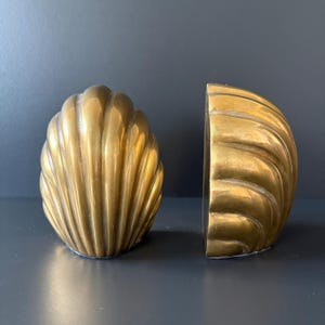 May include: A pair of brass seashell bookends. Each bookend is a half-shell design with detailed ridges, creating a textured surface. The bookends have a warm, golden hue and are designed to stand upright.