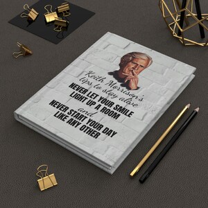 May include: A hardcover journal with a white brick-patterned cover featuring a portrait and text. The text reads "Keith Morrison's tips to stay alive." Gold and black pencils and paper clips are also visible.