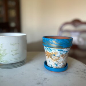 May include: A blue and gold painted terracotta planter with a matching saucer. The planter has a white marbled design with gold accents.