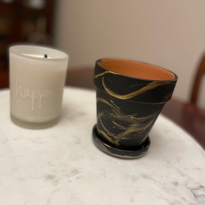 May include: A black and gold painted terracotta planter with a saucer. The planter has a swirl design with gold paint.