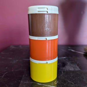Può includere: Una pila di tre contenitori di plastica colorati. Il contenitore superiore è marrone, quello centrale è arancione e quello inferiore è giallo. Ogni contenitore ha un coperchio bianco. I contenitori sono impilati.