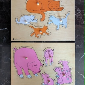 Peut inclure: Deux puzzles en bois à encastrement représentant des familles d'animaux. Le puzzle du haut montre un chat orange et trois chatons blancs et orange. Le puzzle du bas affiche un grand cochon rose et quatre porcelets. Chaque pièce a une petite cheville.