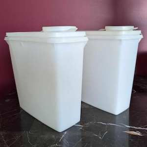 Puede incluir: Dos recipientes blancos y rectangulares para almacenar alimentos con tapas a juego. Los recipientes están hechos de un material plástico translúcido y están sobre una superficie oscura estampada. Las tapas tienen una pequeña abertura circular.