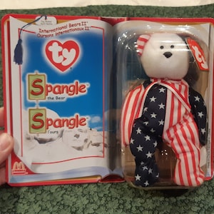 May include: A white beanie baby bear wearing a red, white and blue outfit with stars. The bear is packaged in a plastic case with a Ty tag and a cardboard backing that says "International Bears II" and "Spangle the Bear".