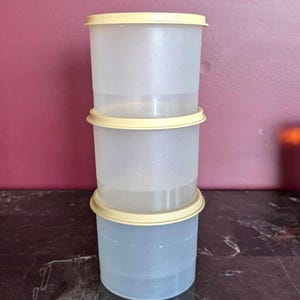 Peut inclure: Trois récipients de stockage alimentaire en plastique transparent empilés les uns sur les autres. Chaque récipient a un couvercle jaune clair. Les récipients sont cylindriques et semblent vides. L'arrière-plan est de couleur marron.