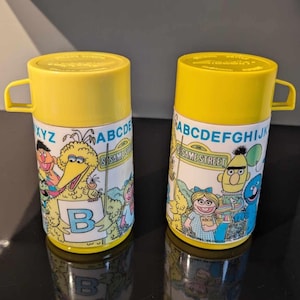 Pode incluir: Duas garrafas térmicas amarelas com personagens da Vila Sésamo. Cada garrafa térmica tem uma tampa e uma alça amarelas. O corpo de cada garrafa térmica é branco com personagens da Vila Sésamo e o alfabeto.