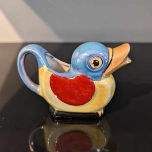 May include: A ceramic bird-shaped creamer. It has a blue head, yellow body, and red breast. The creamer has a handle and a spout, and the beak is orange. The bird is sitting on a black base.