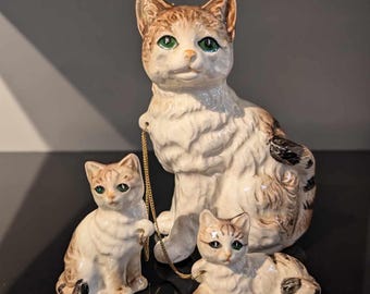 May include: A collection of three ceramic cat ornaments. The largest cat is seated, with two smaller kittens. The cats are white with brown tabby markings and green eyes. A gold-coloured chain connects the large cat to one kitten.