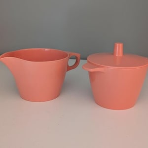 May include: A vintage coral-colored creamer and sugar bowl set. The creamer has a handle and a spout. The sugar bowl has a lid with a small handle. Both pieces are made of a solid, smooth material.