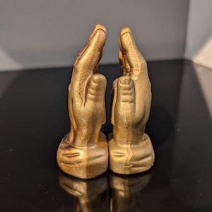 Può includere: Due sculture di mani dorate, palmi rivolti l'uno verso l'altro, in gesto di preghiera o sostegno. Le mani presentano linee e texture dettagliate, su sfondo scuro. Oggetto decorativo.