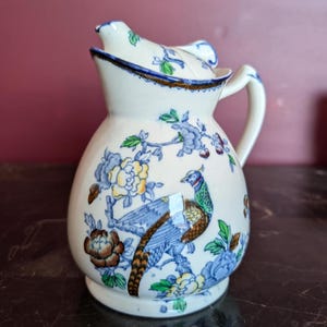 Puede incluir: Jarra de cerámica color crema con tapa, con un diseño detallado de faisán y flores en azul, amarillo y marrón. La jarra tiene un asa curva y un pico con ribete azul. Una pieza decorativa para servir.