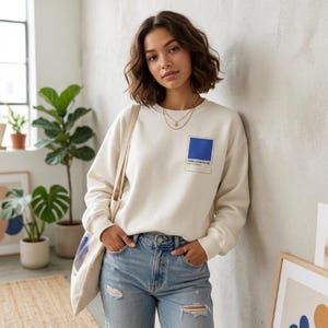 Puede incluir: Sudadera color crema con un gráfico de un cuadrado azul que dice "Cerulean Blue". La sudadera se combina con vaqueros de mezclilla de lavado claro. Una bolsa de lona se lleva sobre el hombro.