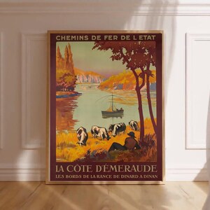 Könnte beinhalten: Ein gerahmtes Vintage-Reiseposter mit dem Text "CHEMINS DE FER DE L ETAT" und "LA COTE D'ÉMERAUDE". Das Poster zeigt eine malerische Landschaft mit einem Fluss, einem Boot, grasenden Kühen und einer sich entspannenden Person. Die Farbpalette umfasst Orange-, Braun- und Grüntöne.