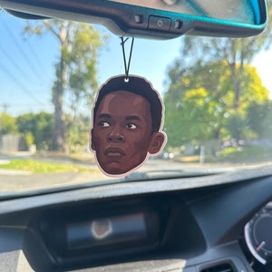 May include: A car air freshener featuring a black and brown illustration of a person's face. The air freshener is hanging from the rearview mirror of a car.